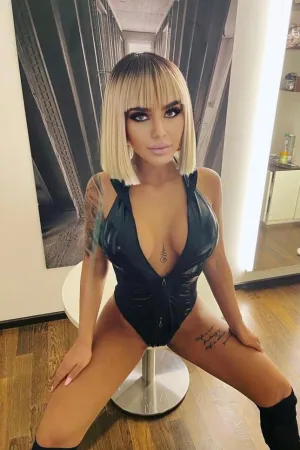 Aida sat on a stool wearing a black bodysuit looking busty